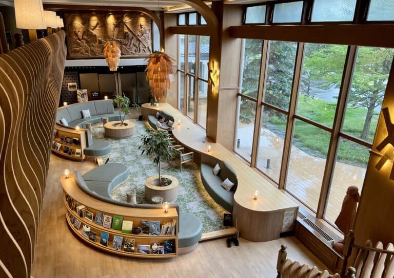 A bright and airy lobby lounge at The Forest Akan with curved seating, large windows, wood-carved wall art, and bookshelves, blending cozy design with natural light.