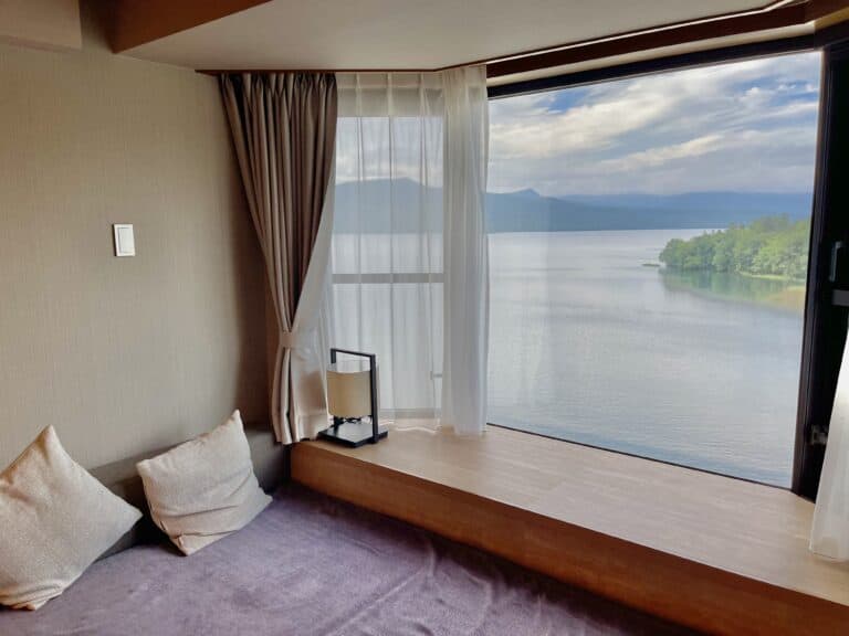A corner room at New Akan Hotel with wide windows revealing a serene lake and mountain view, furnished with cozy pillows and sheer curtains. Perfect for families looking for tranquility at a family friendly onsen hotel in Akan.