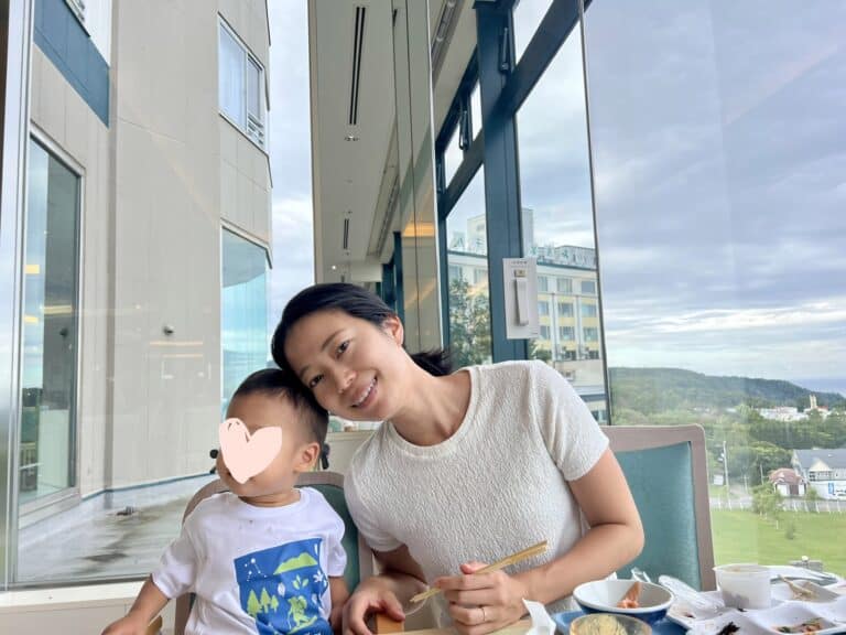 A smiling mother and toddler sit at a dining table inside Shiretoko Daiichi Hotel, with large windows revealing a view of the hotel exterior and surrounding greenery. Various breakfast dishes are laid out in front of them.